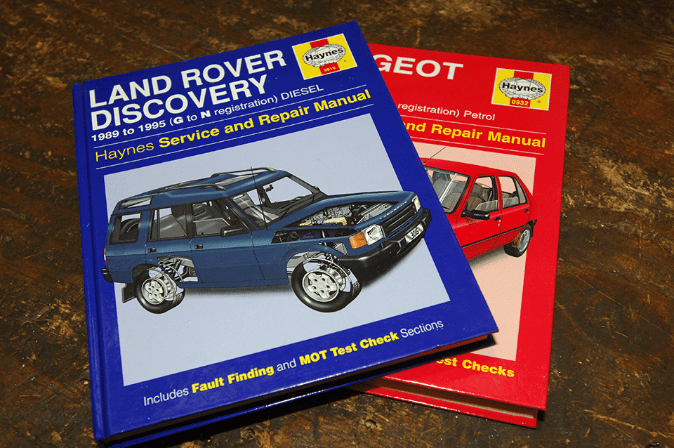 Two Haynes vehicle handbooks lying on a wooden surface; one for a blue Land Rover Discovery diesel and one for a red Peugeot, both showing front car illustrations on the covers.