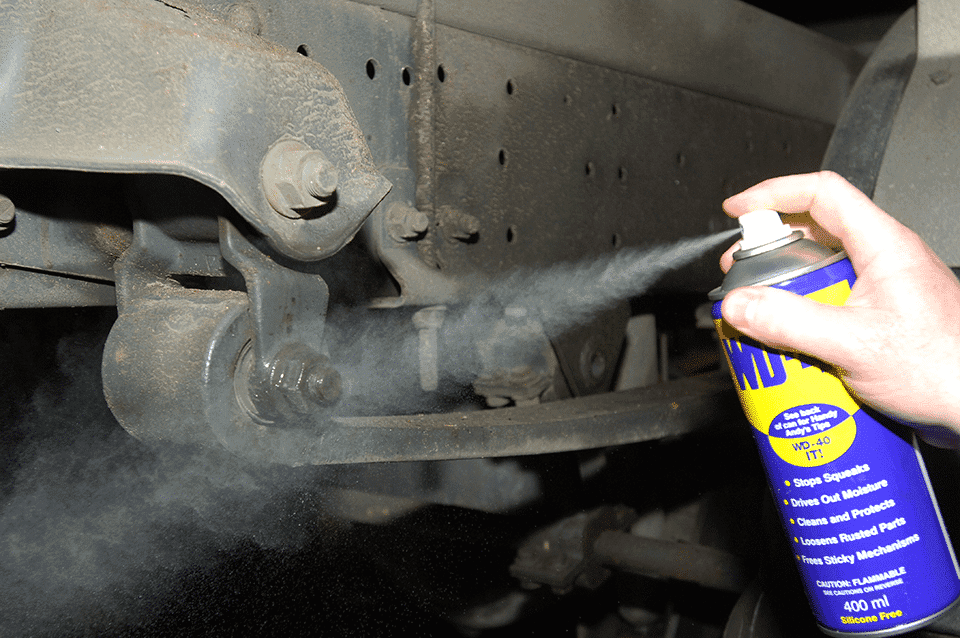 A person applies wd-40 from a tin to a rusty car undercarriage, focusing on a bolt joint to loosen it.