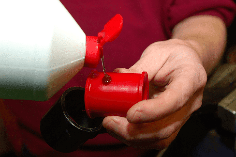 A person clad in a purple shirt pours oil from a white jug into a small red oil can cradled in their hands, demonstrating a routine maintenance or mechanical task.