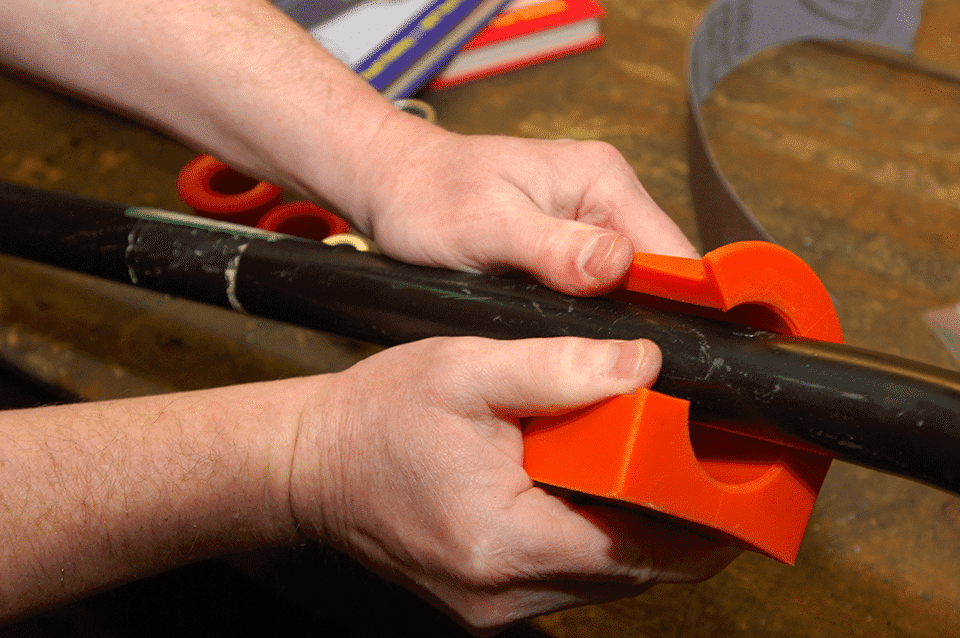 Close-up of two hands using an orange clamp device to cut or secure a thick black cable on a wooden workbench with tools in the background.