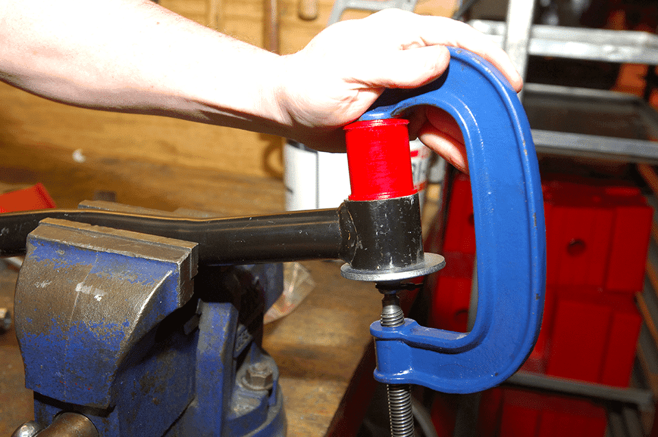 A person's hands utilising a blue vice to securely hold a cylindrical red and black object on a workbench, with tools and boxes visible in the background.