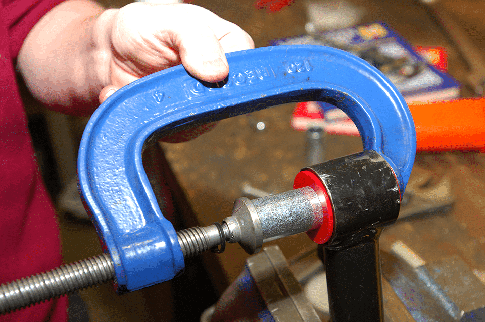 Close-up of a hand tightening a blue G-clamp onto a metal pipe in a workshop, with tools strewn in the background.