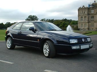 A dark blue Volkswagen Corrado parked on a tarmacked area with a large, imposing manor house and a fountain in the background under a cloudy sky.