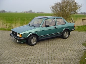 A classic green Volkswagen Golf parked on a cobbled area with a grassy field in the background on a cloudy day.