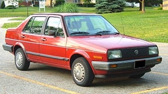 A red Volkswagen Jetta parked on the side of a road with green grass and trees in the background. The car is an older model with a clean and shiny exterior.