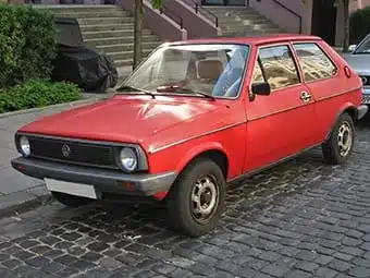A red Volkswagen Golf Mk1 parked on a cobbled street. The compact car features sleek lines and a classic design, displaying a shiny red bodywork and black trim around the windows.