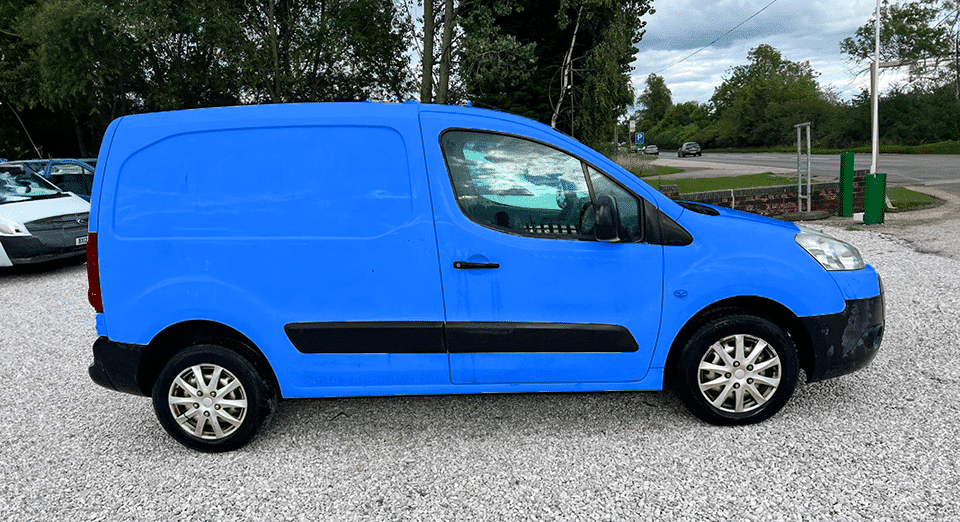 A vivid blue compact cargo van parked on a gravel car park surrounded by trees, with a partly cloudy sky in the backdrop. The van is orientated towards the left with noticeable wear on the front bumper.