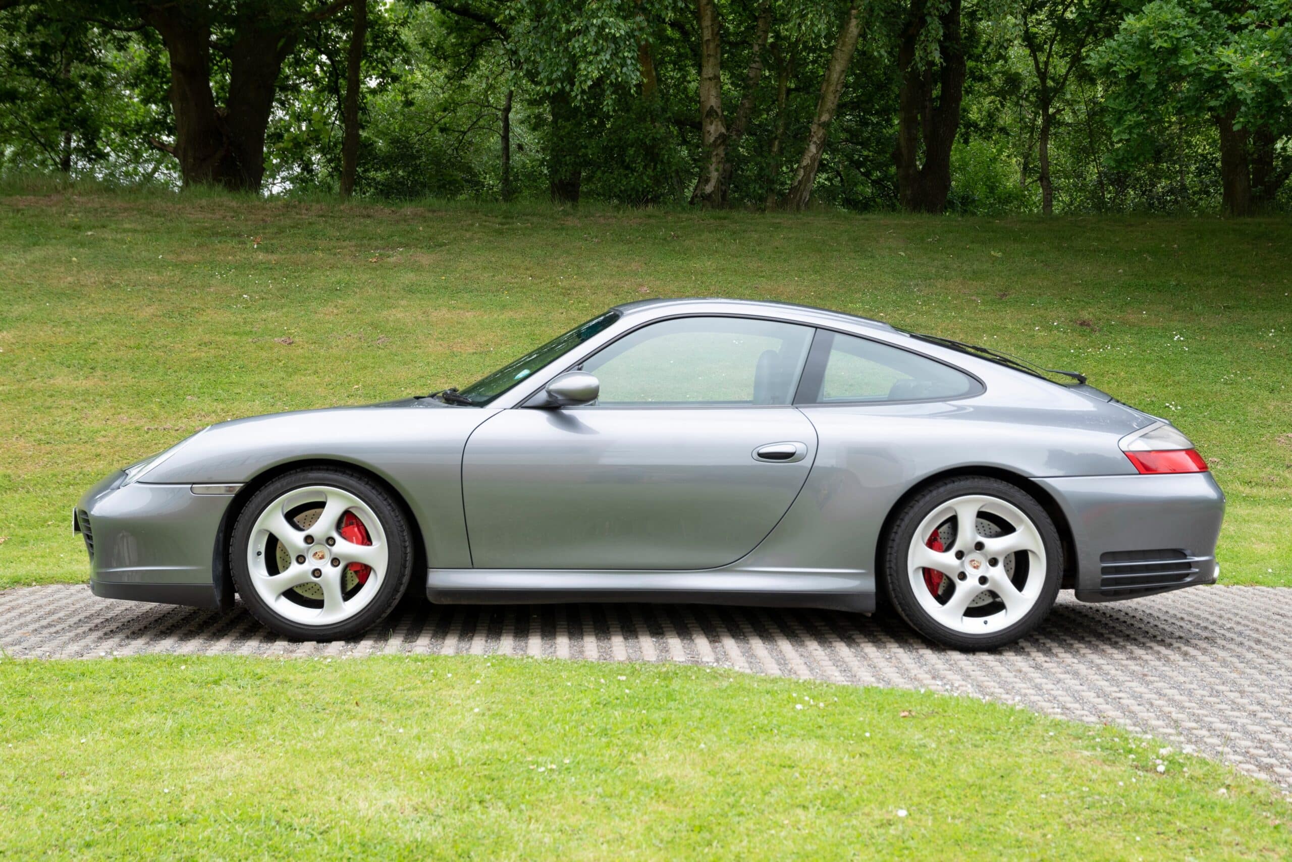 A silver Porsche 911 Carrera parked on a brick pathway beside a grassy area with trees in the background. The car features a sleek design and red brake callipers.