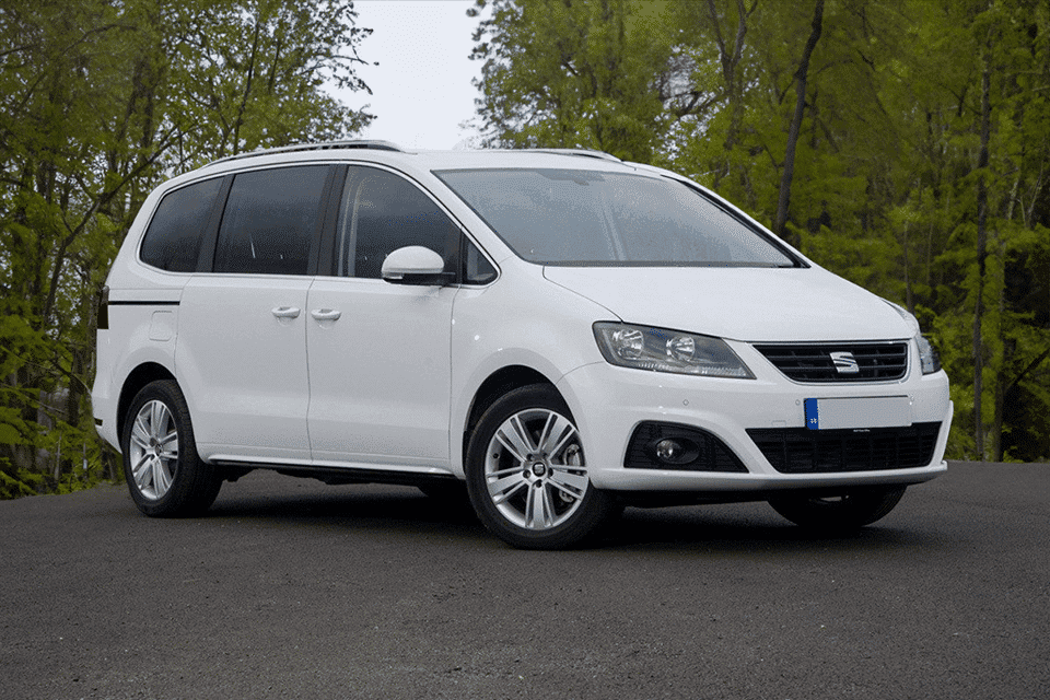 A white Seat Alhambra MPV parked on a tarmac road surrounded by lush green trees. The vehicle boasts a clean, modern design with silver alloys.