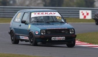 A Volkswagen Golf GTI race car, number 10, speeds on a racecourse, its front tyres lifting slightly off the ground as it corners aggressively. The car is adorned with various sponsor logos.