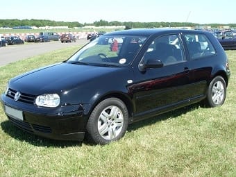 A black Volkswagen Golf parked on a grassy field with a clear blue sky overhead, surrounded by multiple other parked cars in the background.