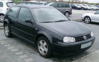 A black Volkswagen Golf parked in a car park, boasting sleek lines, gleaming wheels, and lightly tinted windows beneath a cloud-filled sky.