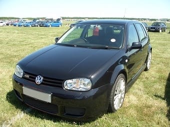 A black Volkswagen Golf parked on a grassy field on a sunny day, with other cars in the background and a clear blue sky.