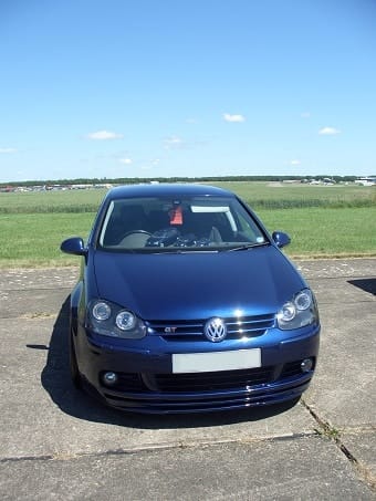 A blue Volkswagen Golf parked on a broad, open airfield under a clear blue sky. The car is facing the camera, showcasing its front design and headlights.