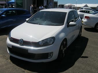 A white Volkswagen Golf GTI parked outdoors at a car event with a crowd and other vehicles in the background under a clear sky.
