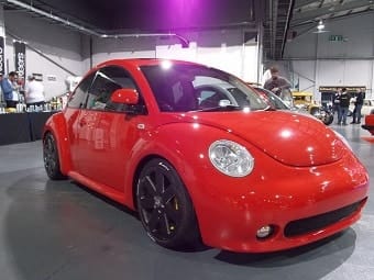 A shiny red Volkswagen Beetle exhibited at an indoor motor show, featuring a sleek design and large alloy wheels, with people in the background.