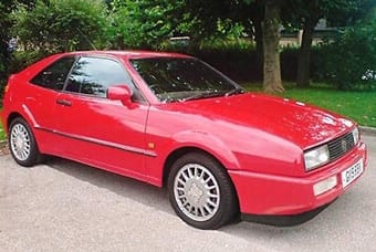 A red vintage sports car parked outside, featuring a sleek design with pop-up headlights and five-spoke alloy wheels. Surrounded by foliage in a tranquil setting.