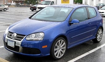 A blue Volkswagen Golf parked in a damp car park on a rainy day, showing raindrops on the car surface.