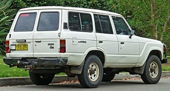 A white, vintage Toyota Land Cruiser parked on a street, with a backdrop of lush greenery. The 4x4 looks robust, showing some signs of wear on its body.