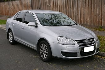 A silver Volkswagen saloon parked on a road, slightly dusty, with a wooden fence in the background. Number plate is obscured.