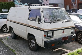 An old white Mitsubishi van parked on a street, featuring a two-tone paint scheme with a beige lower half, embellished with multiple stickers on its window.