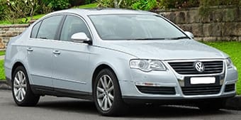 Silver Volkswagen Passat parked on a road next to a stone wall, partially hidden by foliage.