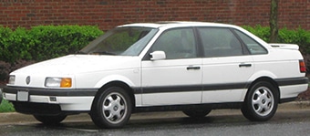 A white four-door saloon Volkswagen Passat parked by the roadside on a wet day, featuring a streamlined body and distinctive wheel design.